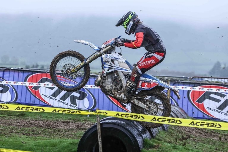 Endurocross racer launching over a row of tires on the track. He's wearing Risk Racing riding gear and his bike is equipped with Plews Tyres.