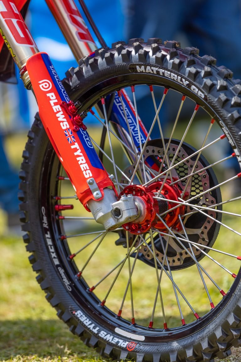 close up of a front tire and front forks of a dirt bike in an outdoor setting. The tire is a Plews Tyre Matterly GP and the fork covers are dawning a Plews Tyres sticker