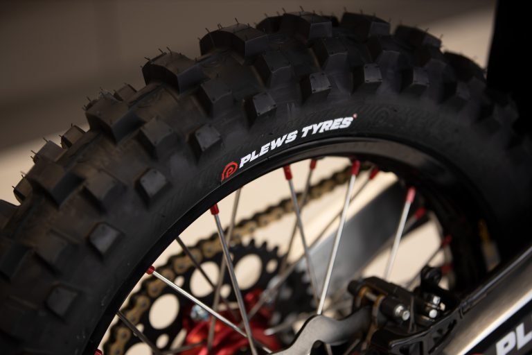 Super close up on a rear dirt bike tire with the Plews Tyres logo prominently displayed in white against the black rubber.